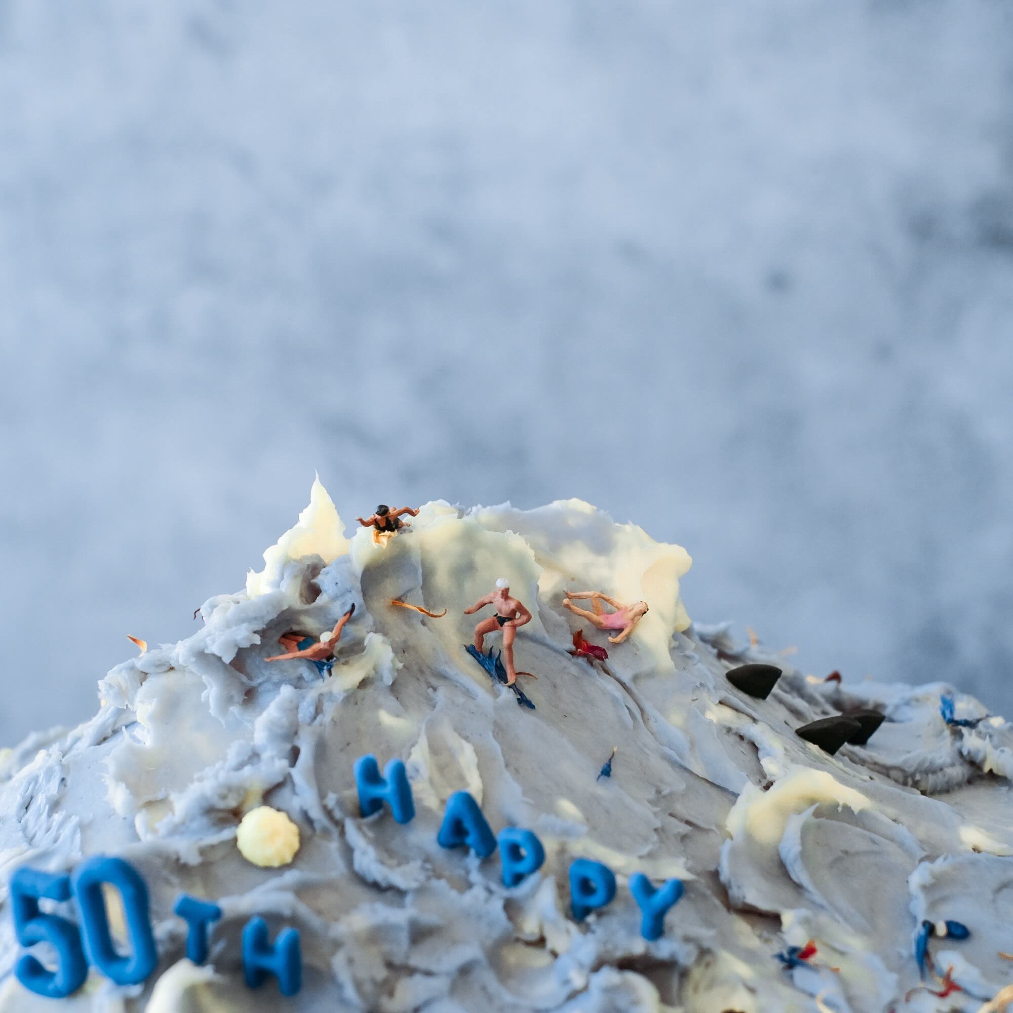 Close-up of beach-themed cake top with sculpted buttercream waves and tiny swimmer figurines, capturing an aerial ocean view effect.