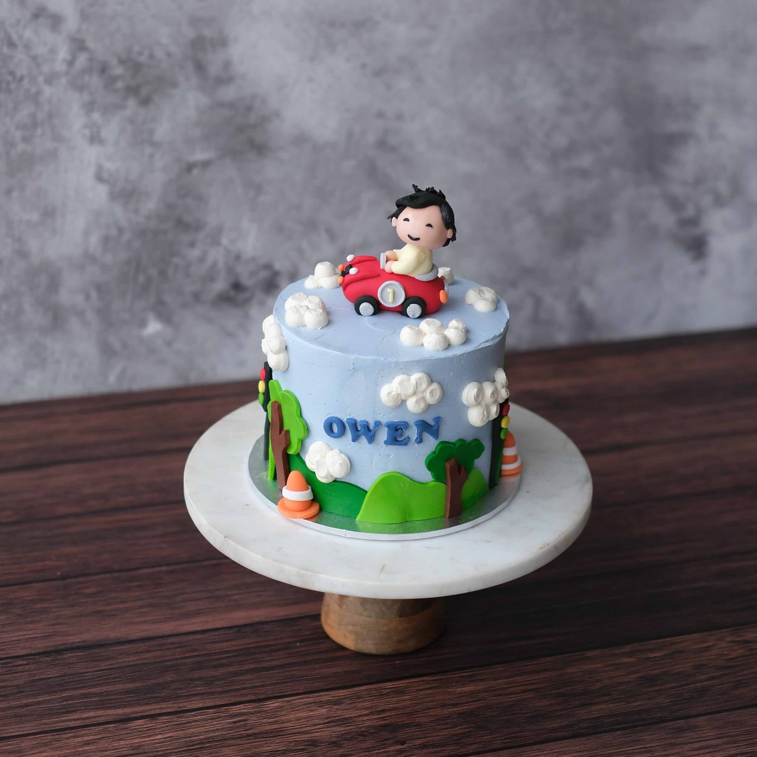 First birthday cake with pastel blue buttercream, fondant boy in a red car, clouds and personalised name, styled for a playful one-year celebration.