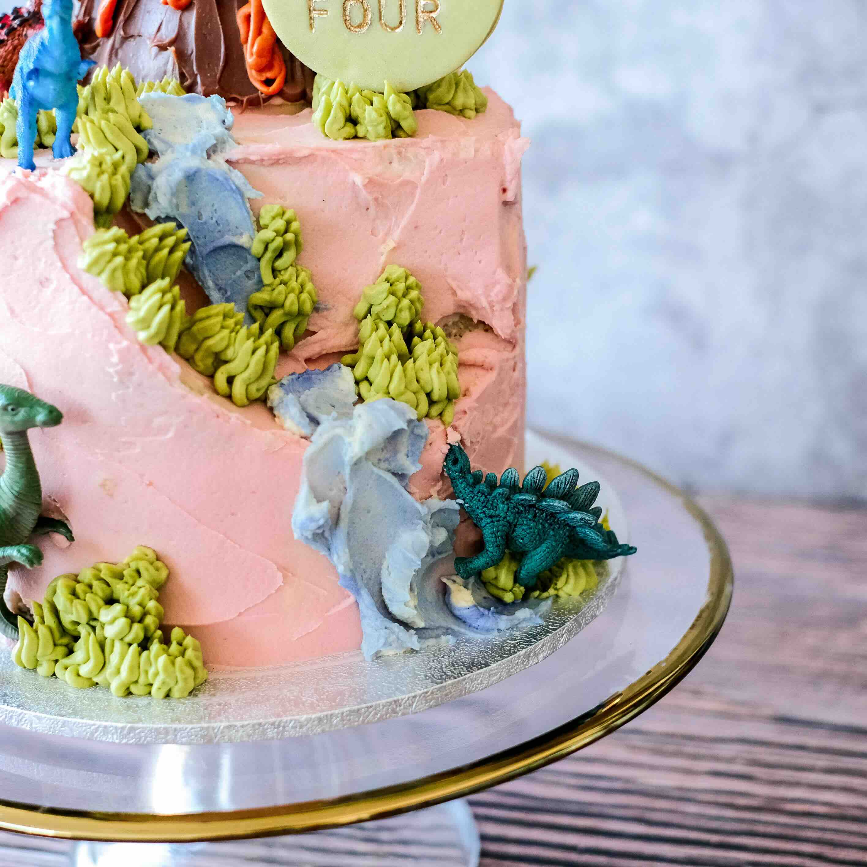 Dinosaur-themed birthday cake with pink frosting, greenery, and small dinosaur figurines.