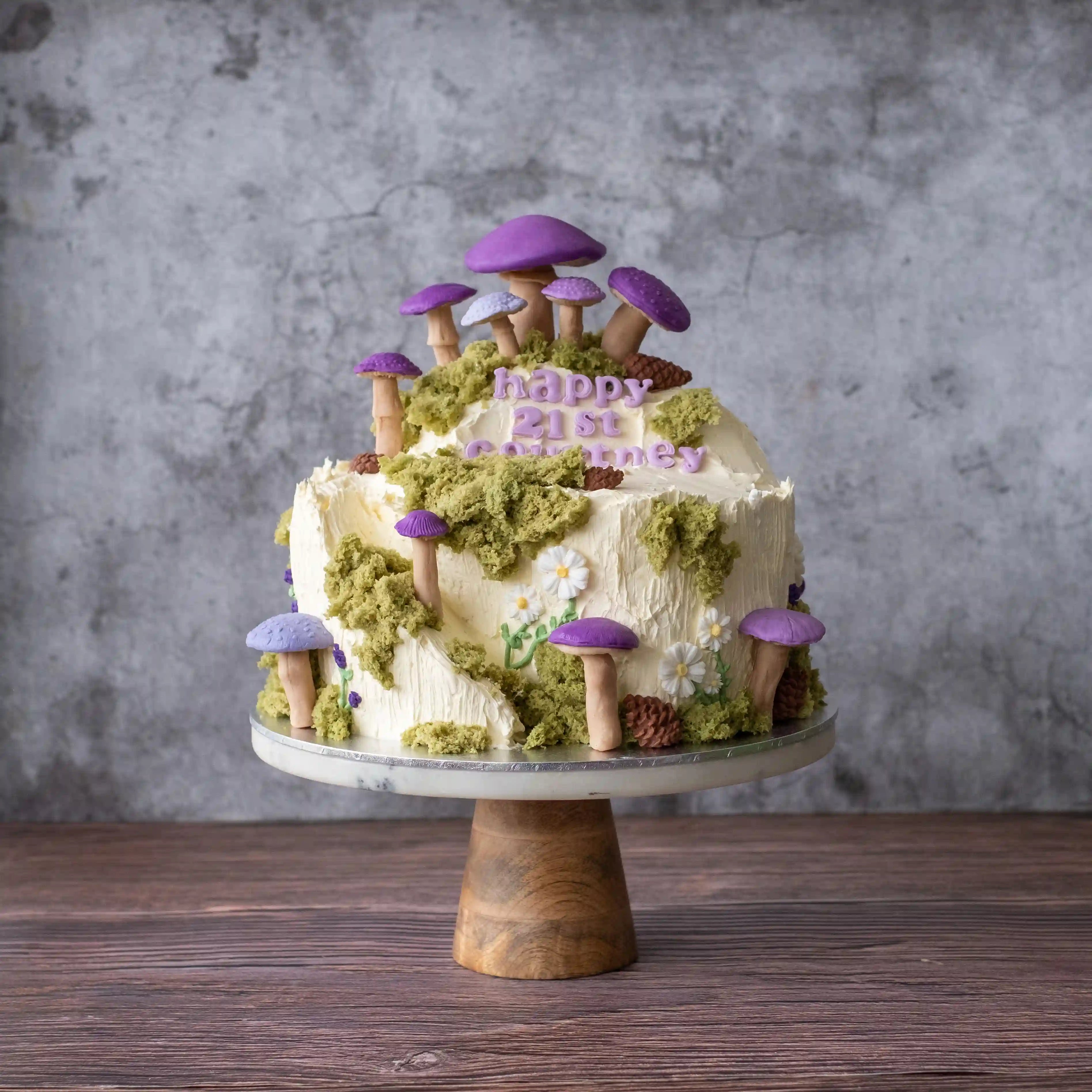 Woodland-inspired celebration cake decorated with purple fondant mushrooms, matcha sponge moss, buttercream florals and a rustic forest-style finish.