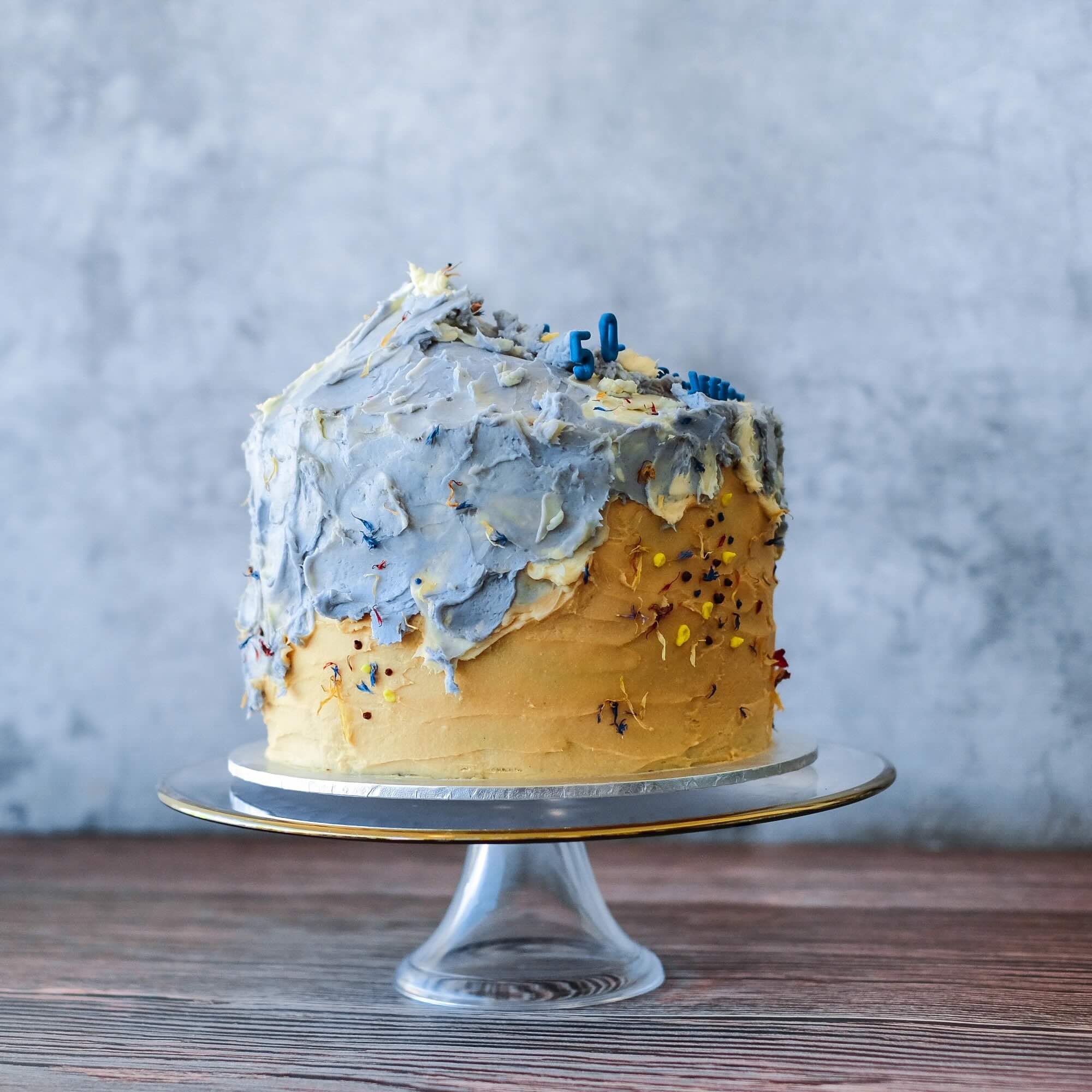 Beach-inspired cake with sandy buttercream tones, soft blue wave-like frosting on top, and scattered edible petals on a glass cake stand.