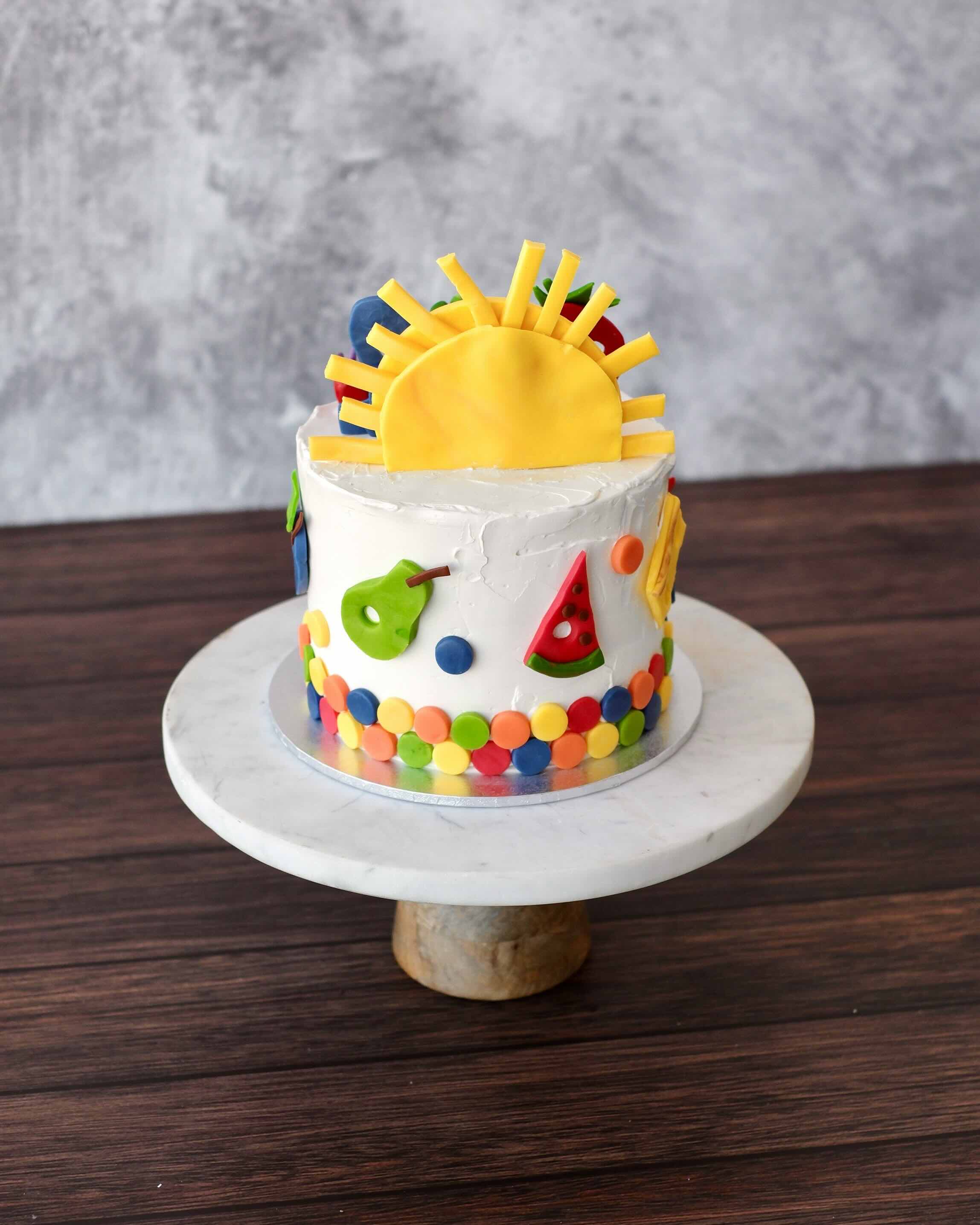 Hungry Caterpillar themed birthday cake with colourful fondant caterpillar, fruits, sun on white buttercream. Back View.