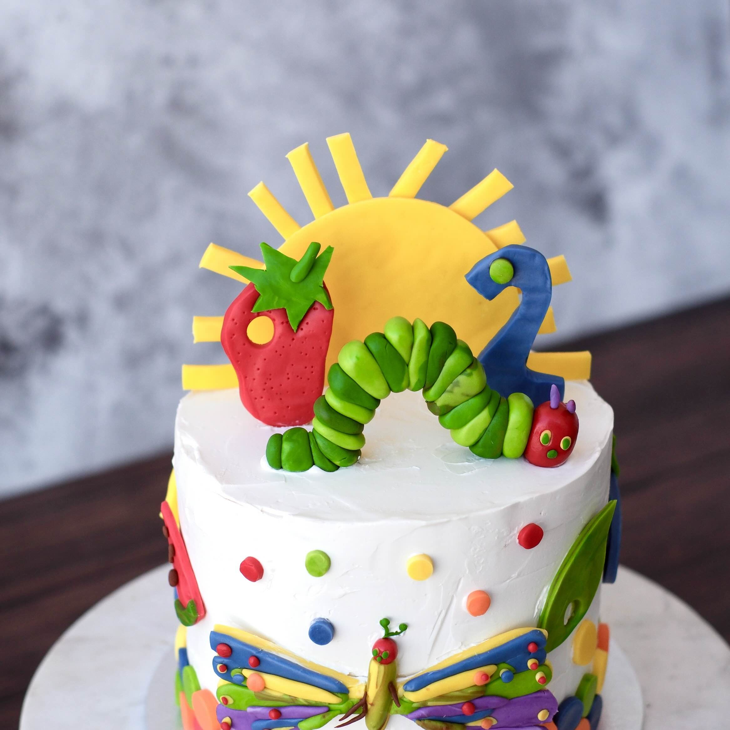 Hungry Caterpillar themed birthday cake with colourful fondant caterpillar, strawberry, sun and bright playful details on white buttercream.